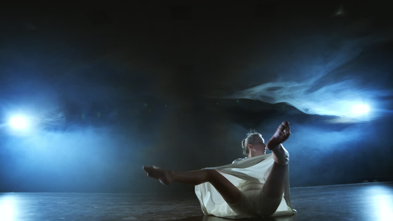 Modern dance girl in a white dress dances a modern ballet jumps on the stage with smoke in the blue spotlights.