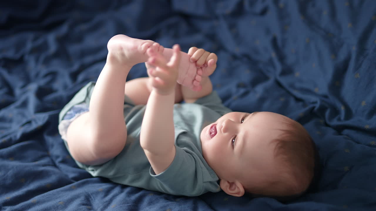 Adorable Caucasian toddler lies on his back on the bed. Beautiful child plays with his feet pulling them to mouth.