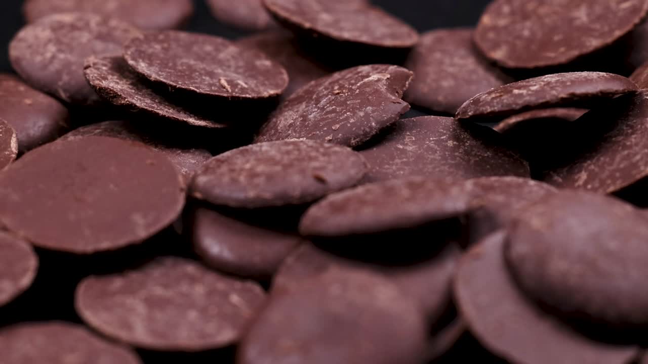 Chocolate discs cascade and accumulate on a dark surface, creating a textured pile.