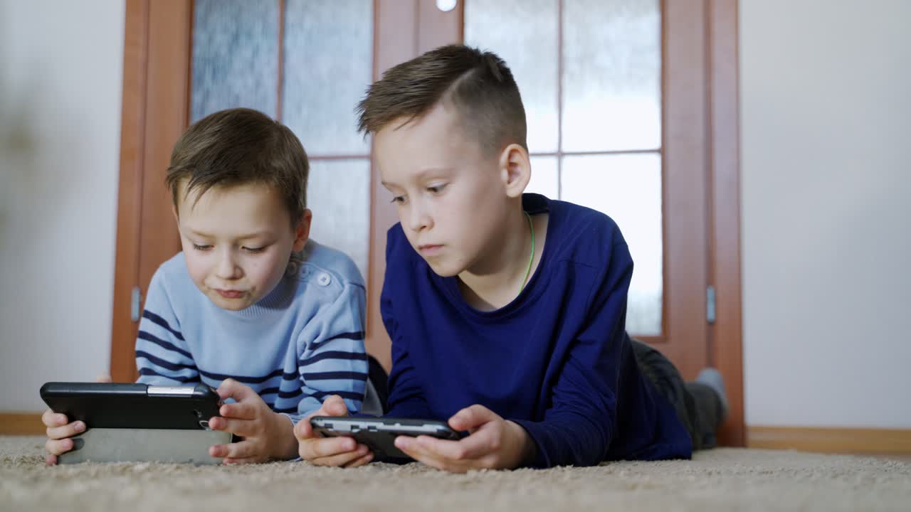 Cute boys lying on the floor in the living-room and playing on modern devices with excited faces. Children enjoying with their gadgets and showing interesting moments each other at home.