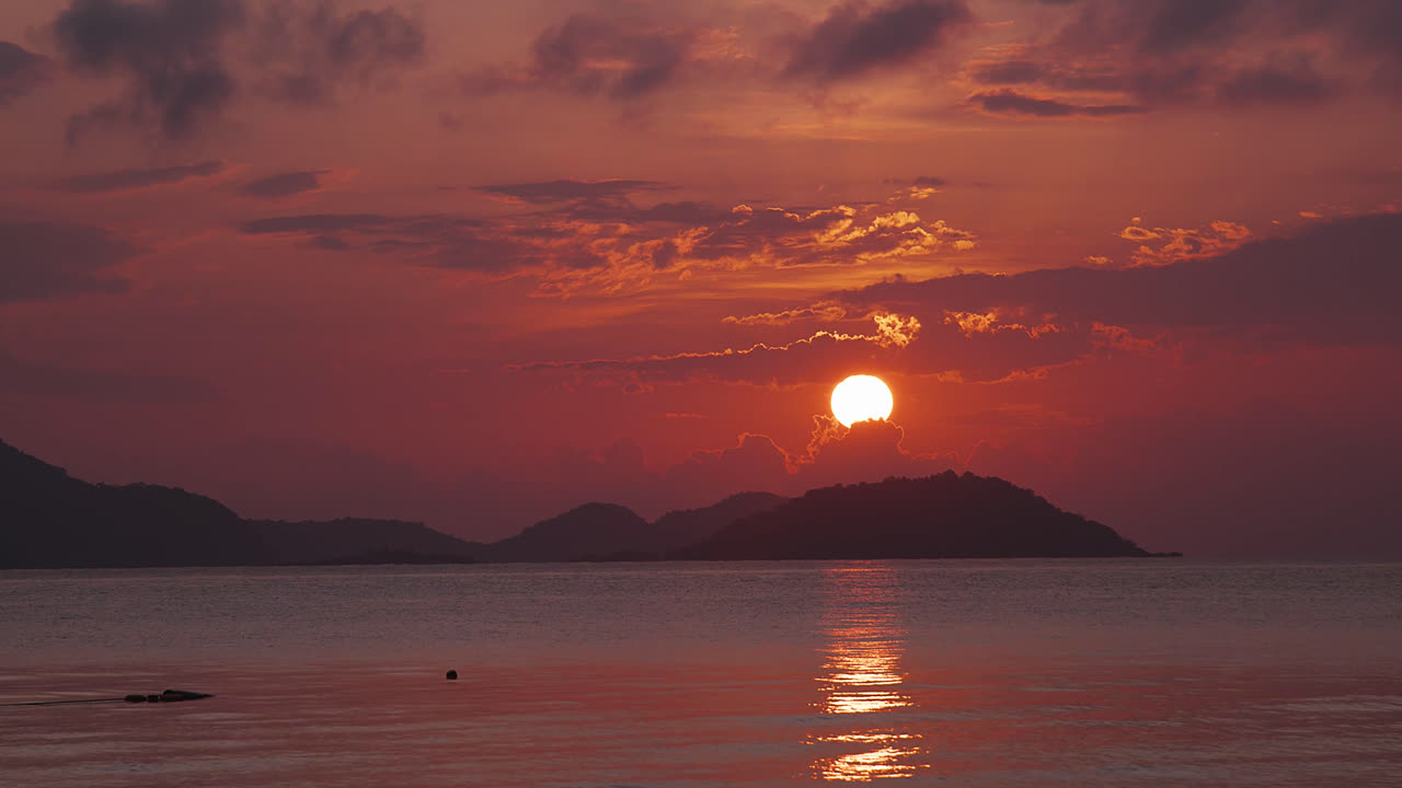 壮大な日の出。穏やかな海にはさざ波が立ち、背景には島が浮かぶ。オレンジ色の太陽が黄金色の光を投げかけ、紫色の雲を縁取る景色。