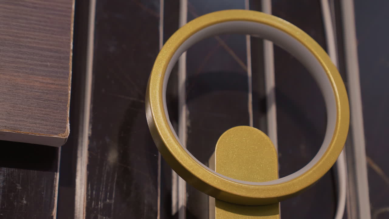 Gold ring light closeup on black painting, bright LED halo and reflective glass, textured wood slats and glossy stripes, warm amber glow creating cozy cinematic ambiance, detailed metal finish