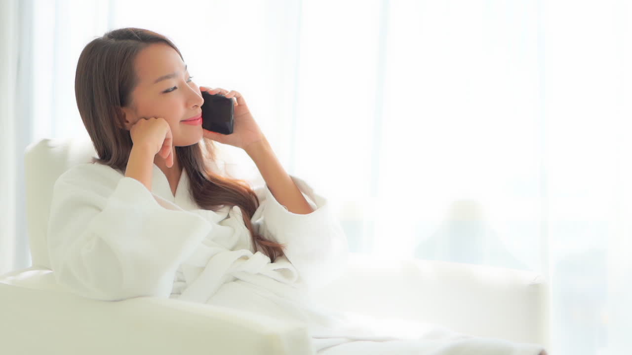 테리 옷을 입은 목욕 가운을 입고 편안한 의자에 앉아 휴대폰으로 이야기하는 예쁜 여성의 클로즈업