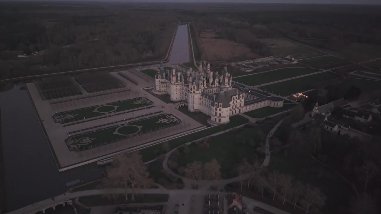 Une vue aérienne montre le canal et les jardins à la française du Château de Chambord s'illuminant doucement au crépuscule