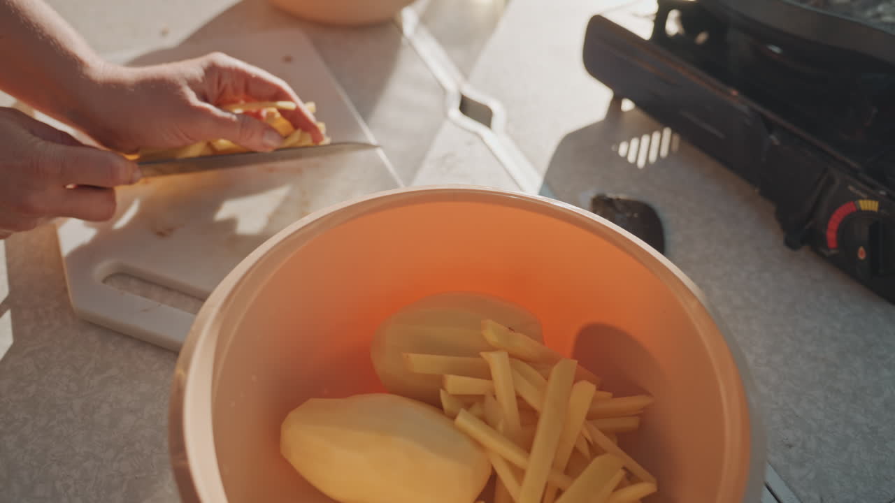 Hands loading fries into orange bowl near stove, mushrooms in separate bowl and portable burner beside, sunlight on table, prepping potatoes for frying, friendly campsite kitchen vibe