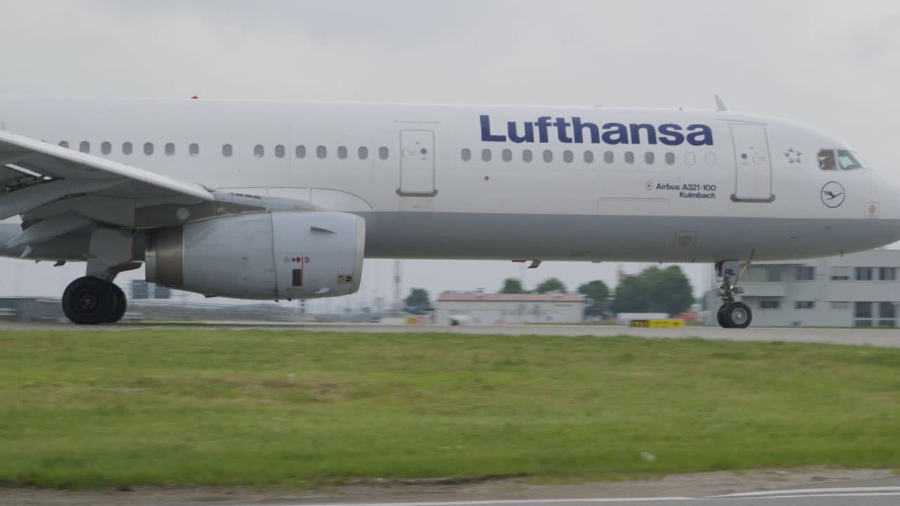 Medium wide shot of an Lufthansa airplane rolling on the airfield to the assigned terminal. Camera pans with the plane. Cloudy day