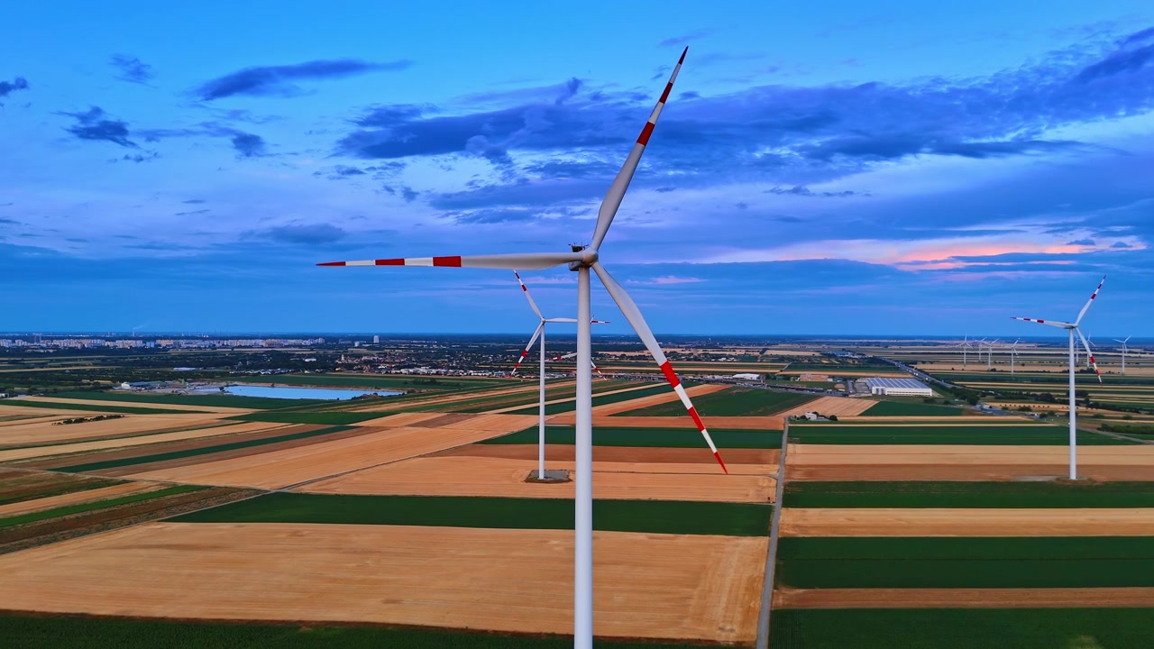 Øko-vindgeneratorer på mørke lappetæppe-marker i skumringen. Luftpanorama af flere elektriske turbiner over landområder under dybblå himmel med orange skær.