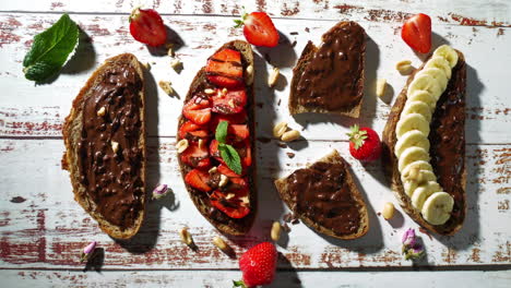 overhead shot of various slices of toast with chocolate spread topped with bananas, strawberries, and nuts, as a hand reaches in to take a slice. rustic white wooden table background.