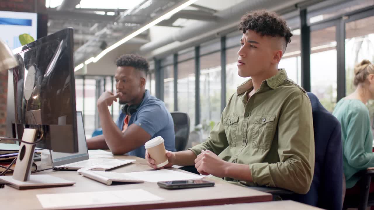 Two diverse male casual business colleagues working at computers and drinking coffee, slow motion