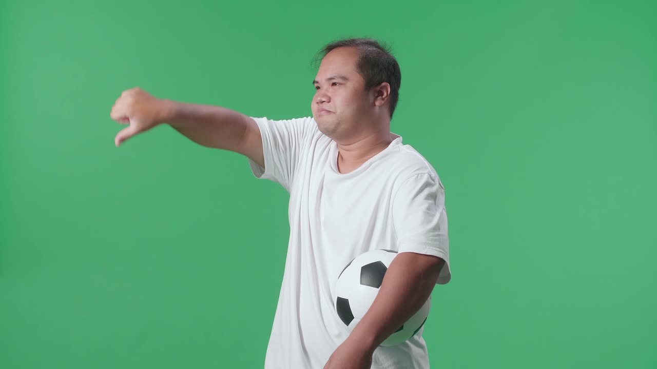 푸른 스크린 배경에서 축구를 응원하는 동안 패자들을 조롱하는 몸짓으로 엄지손가락을 내려놓은 공을 가진 아시아 남자의 측면 시각