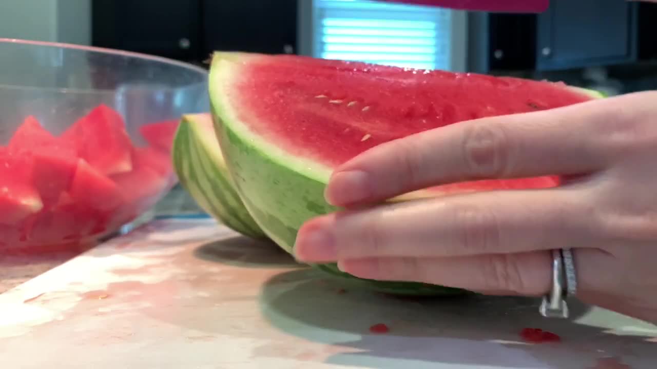 Hand slicing half a watermelon into many triangular pieces with a pink ceramic kitchen knife. 4K closeup video