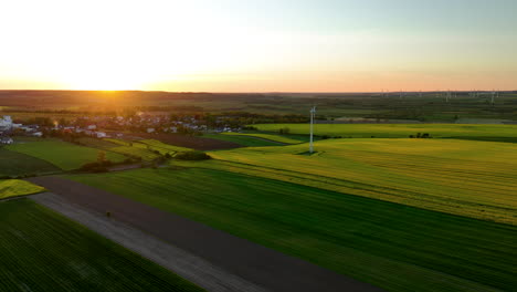 Vista-Aérea-De-Un-Pueblo-Rodeado-De-Campos-Verdes-Al-Atardecer-Con-Turbinas-Eólicas-Al-Fondo