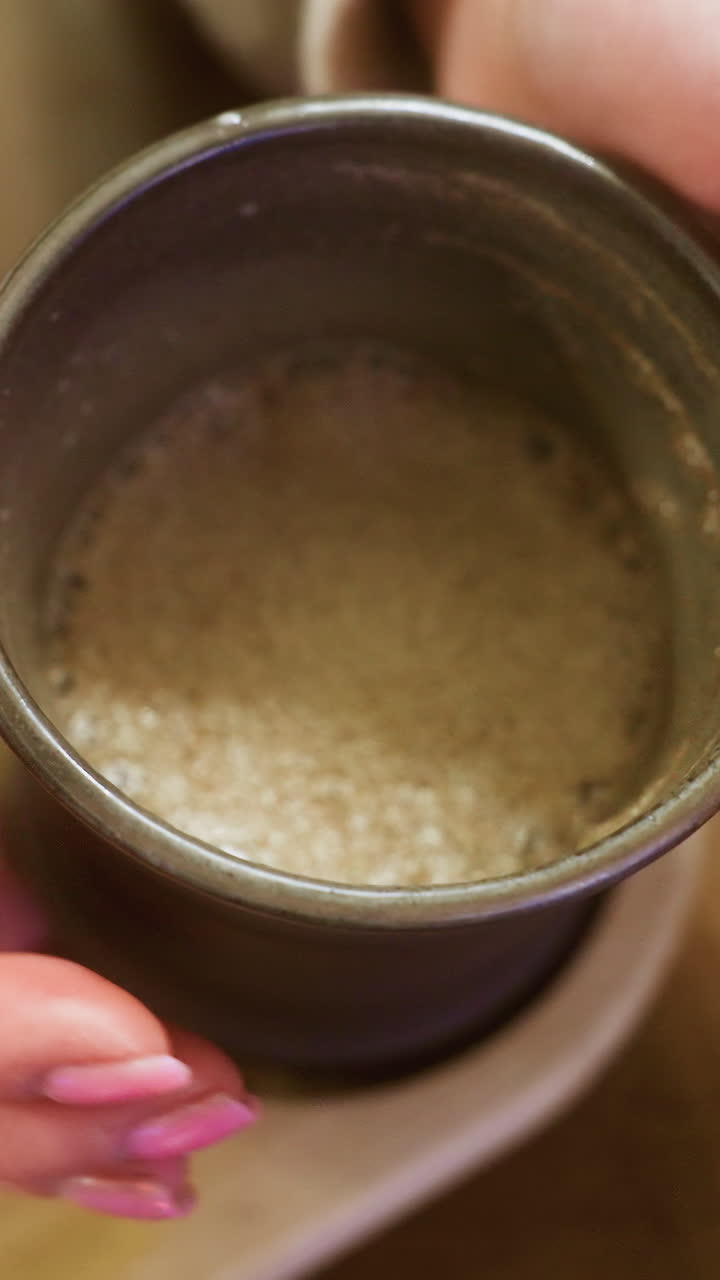 Close-up of lady's hand in brown shearling jacket gently lifting cup of coffee at cozy cafe table. Soft lighting creates inviting and warm atmosphere perfect for peaceful moments