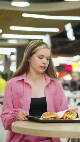 dama de vestido rosa se sienta, se prepara para comer su desayuno con una expresión seria en su cara, bandeja con café, hamburguesa y papas fritas en la mesa, fondo borroso con iluminación bokeh
