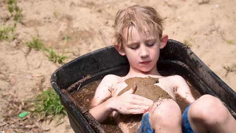 young-boy-covering-chest-in-mud-in-wheelbarrow-filled-with-water