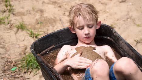 young boy covering chest in mud in wheelbarrow filled with water