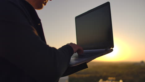 un programador con una computadora portátil se sienta en el techo y escribe código al atardecer. trabajo remoto freelance. libertad para trabajar. escribir en un teclado al atarrear el sol con una vista de la ciudad.