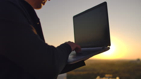 a programmer with a laptop sits on the roof and writes code at sunset. remote work freelancer. freedom to work. typing on a keyboard at sunset with a view of the city.