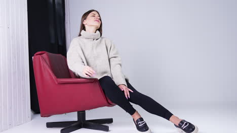 young beautiful long-haired brunette wearing warm sweater, tight pants and stylish shoes sitting in red armchair indoors. model posing in the studio.