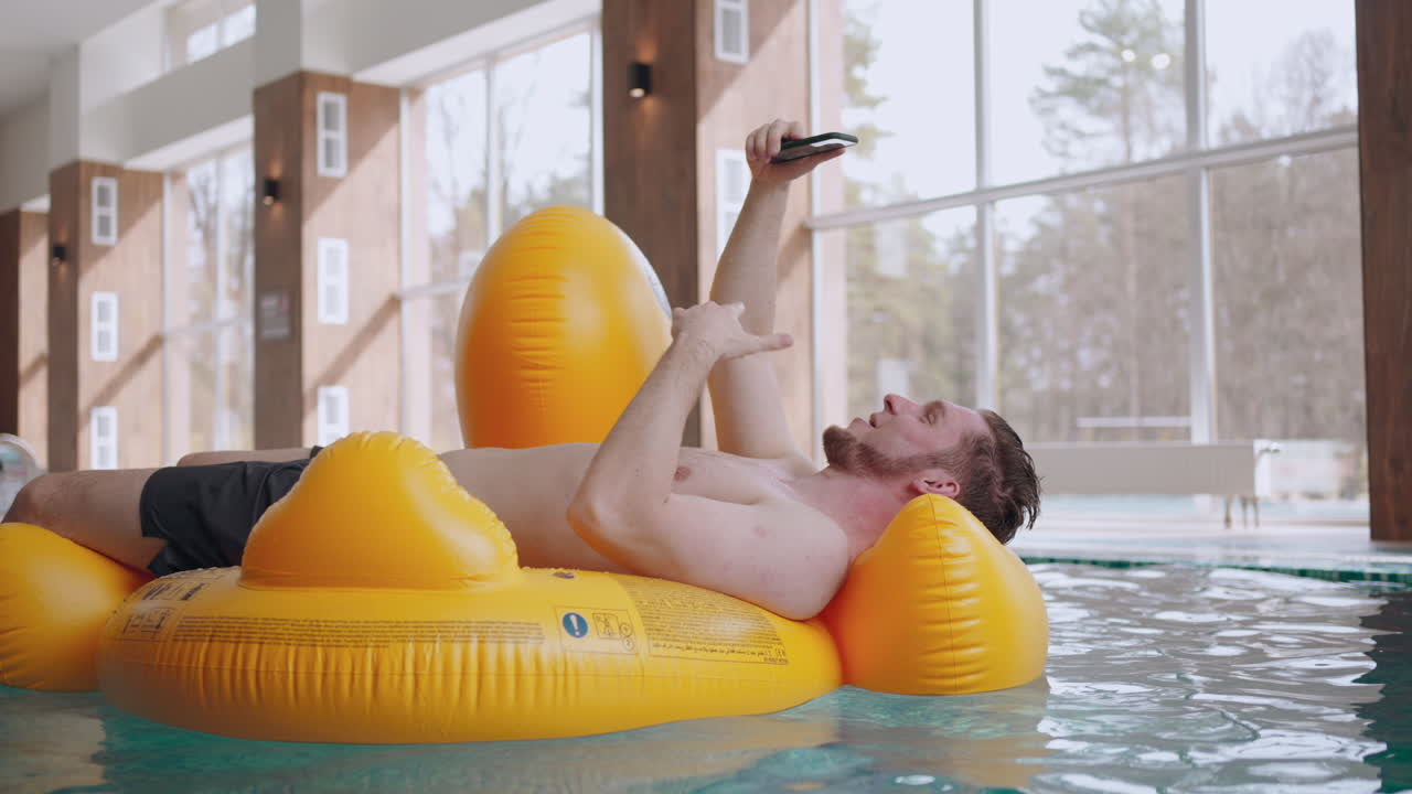happy man is lying on inflatable circle in swimming pool relaxing and talking by video call in smartphone