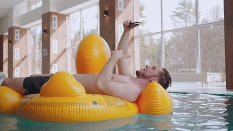 Ein-Glücklicher-Mann-Liegt-Auf-Einem-Aufblasbaren-Kreis-Im-Schwimmbad-Und-Entspannt-Sich-Und-Spricht-Per-Videoanruf-Im-Smartphone