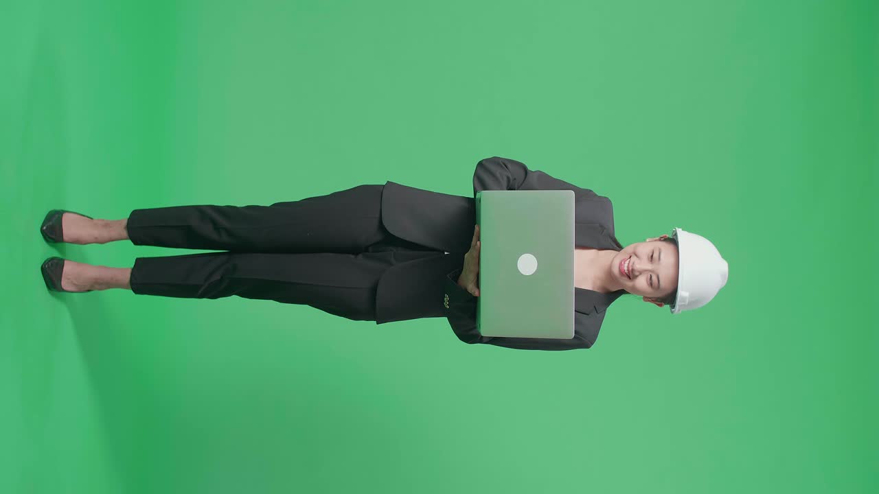 Full Body Of Smiling Asian Female Engineer With Safety Helmet Using A Laptop In The Green Screen Studio