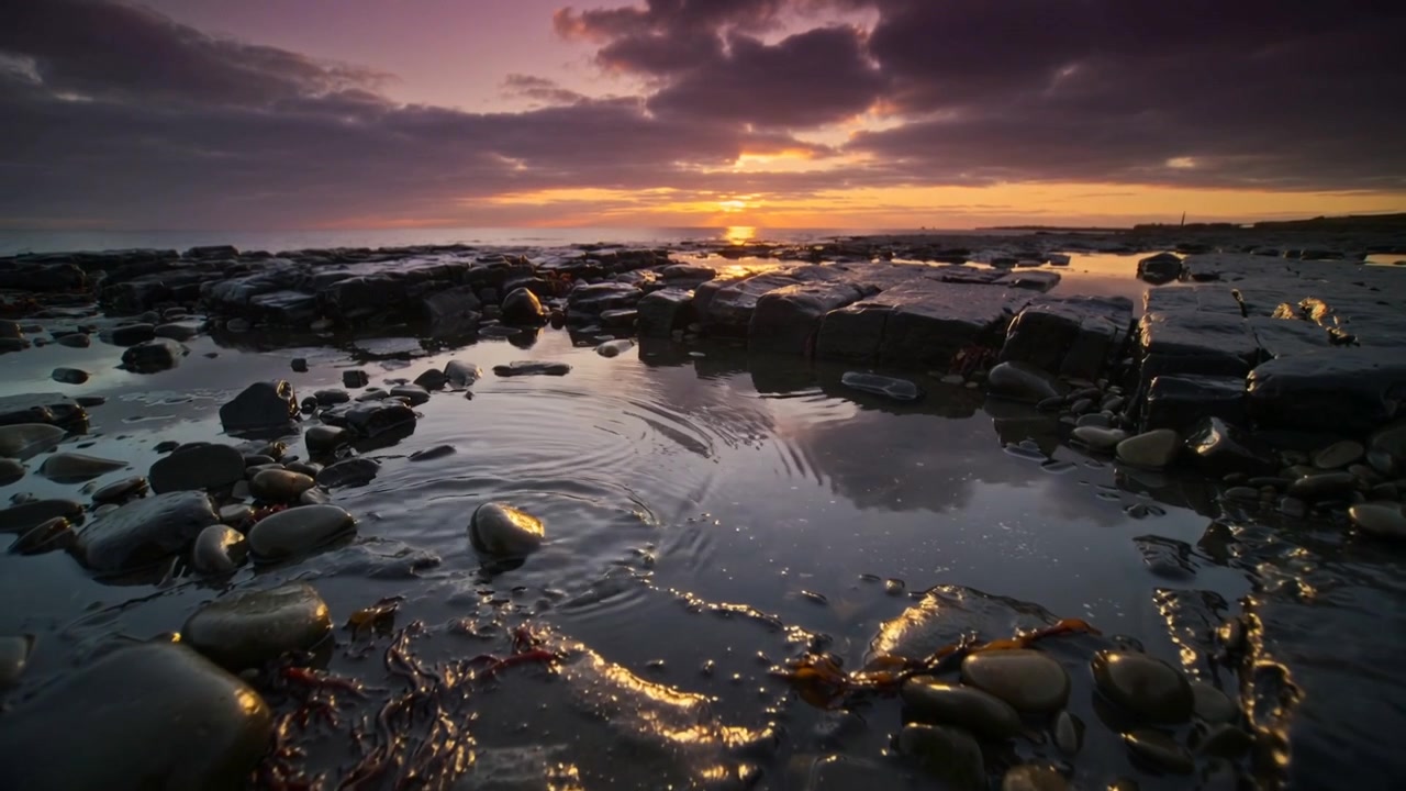 Pozza di marea poco profonda che reagisce a una piccola perturbazione, inviando increspature sulla riva ciottolosa al tramonto