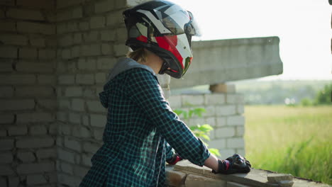 a lady in a helmet stands by the window of an old building, resting her hands on the sill, gazing thoughtfully at the field beyond, with a serene rural landscape stretching in the distance