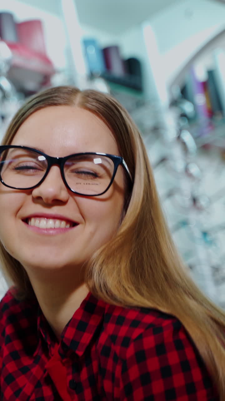 Smiling girl buying eyeglasses in the shop. Attractive young woman trying on new glasses and posing on camera at optics store. Vertical video