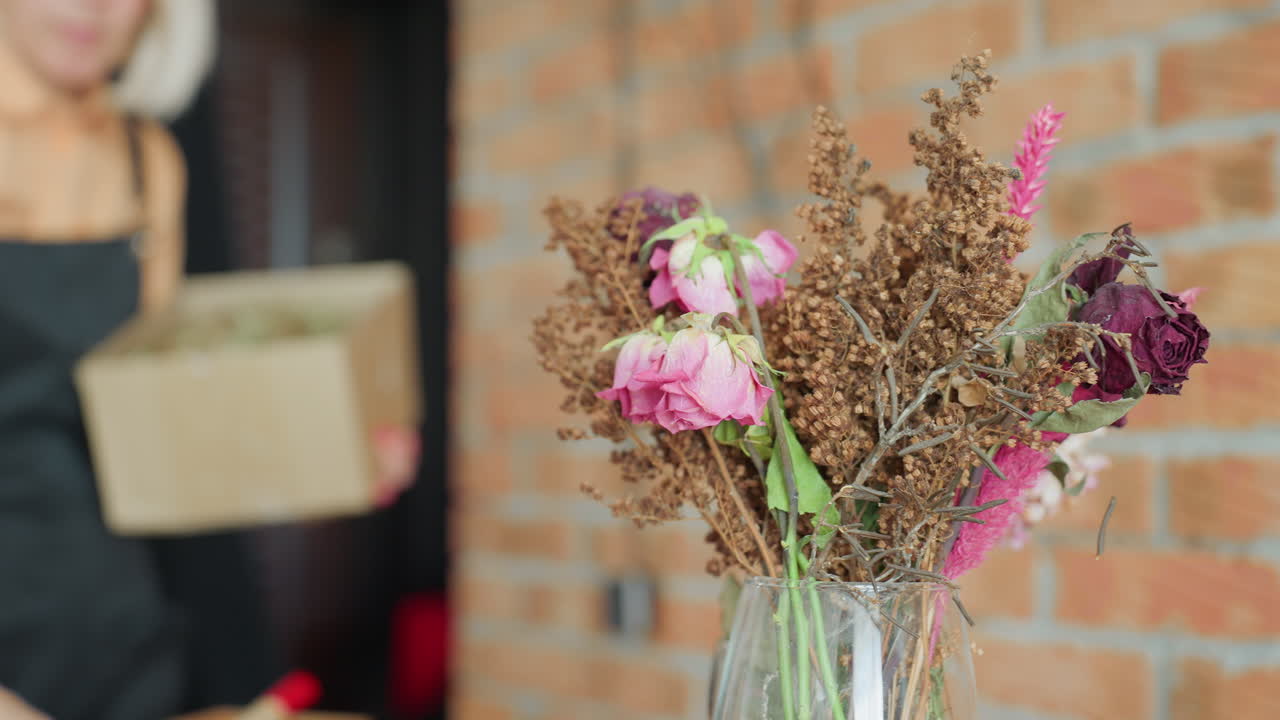 Florist in black apron working with dried flowers arranging bouquet with wilted roses, pink blossoms, in creative studio interior with brick wall background, preparing floral composition for decor