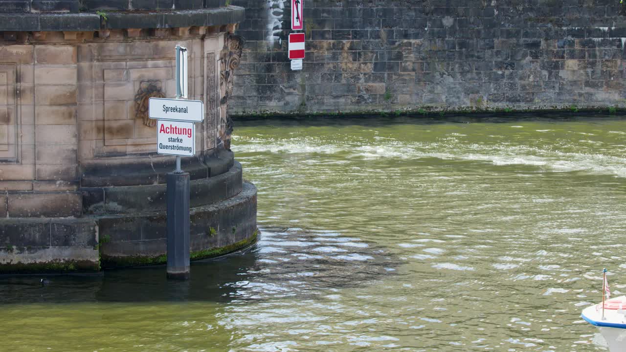 Tourist boats move past stone wall and warning sign on sunny Berlin canal, steady camera