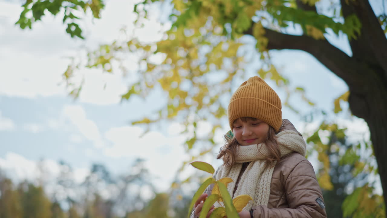 back view close up of kid admiring long yellow autumn leaf while standing on leaf covered path under soft cloudy sky wearing mustard beanie beige jacket and knitted scarf in quiet park
