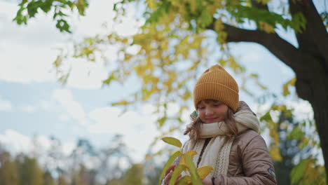 back view close up of kid admiring long yellow autumn leaf while standing on leaf covered path under soft cloudy sky wearing mustard beanie beige jacket and knitted scarf in quiet park