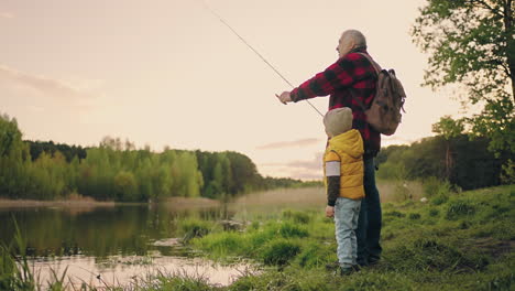 old man and little boy are fishing on shore of picturesque lake in forest grandfather and grandson