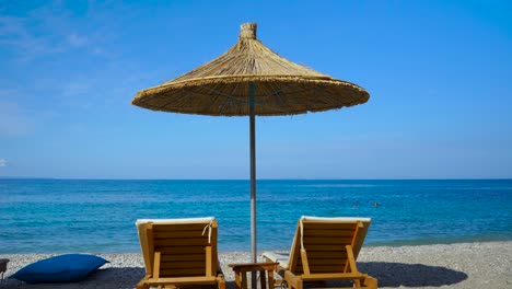 Straw-umbrella-and-sunbeds-on-peaceful-beach-with-blue-sea-and-sky-background,-summer-vacation