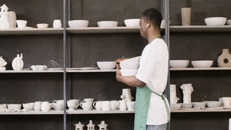 commis masculin avec écouteurs sans fil stockant un bol en céramique sur l'étagère du magasin de poterie 2