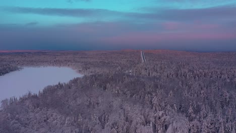 Aerial-shot-flying-forwards-down-towards-a-snowy-forest-and-lake-with-pink-sunrise-skies