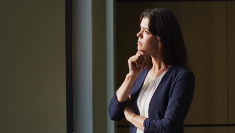 Porträt-Einer-Nachdenklichen-Kaukasischen-Geschäftsfrau-Mit-Braunen-Haaren,-Die-Am-Fenster-In-Einem-Modernen-Büro-Steht