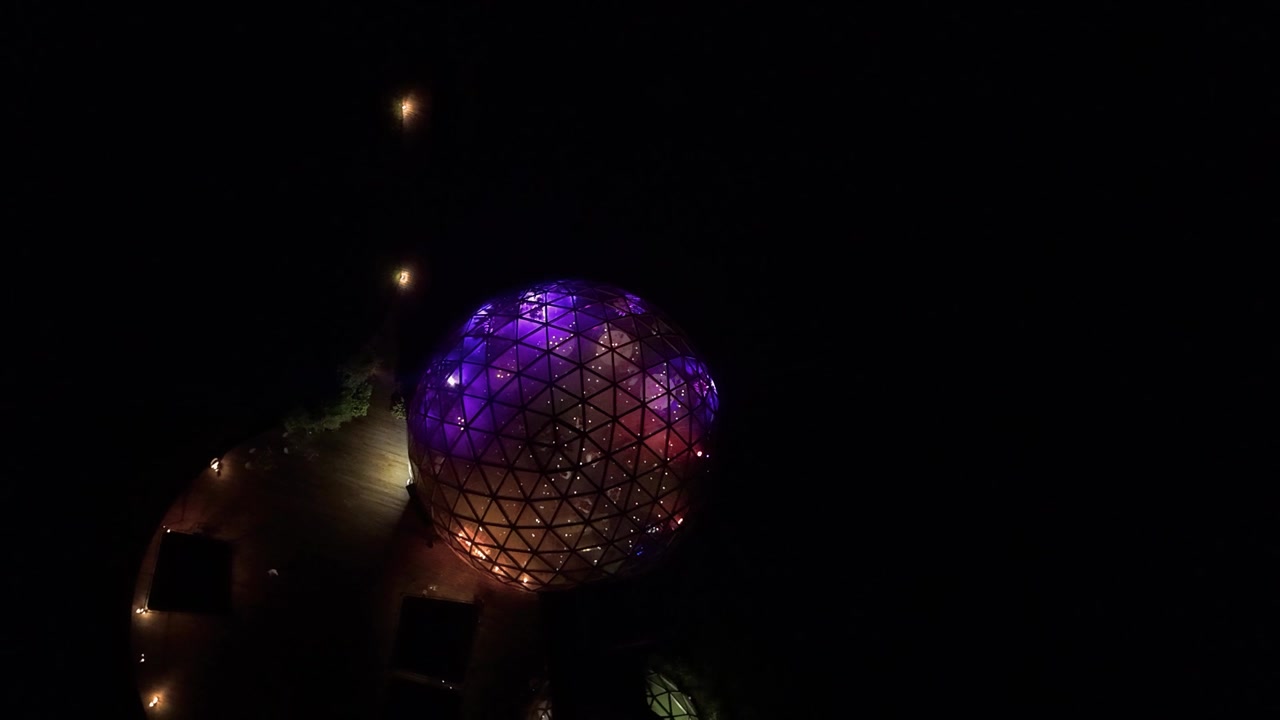 Splendido video aereo con drone che mostra una cupola geodetica luminosa illuminata da vivaci luci viola e arancioni contro un paesaggio notturno scuro, con architettura moderna e contrasti drammatici