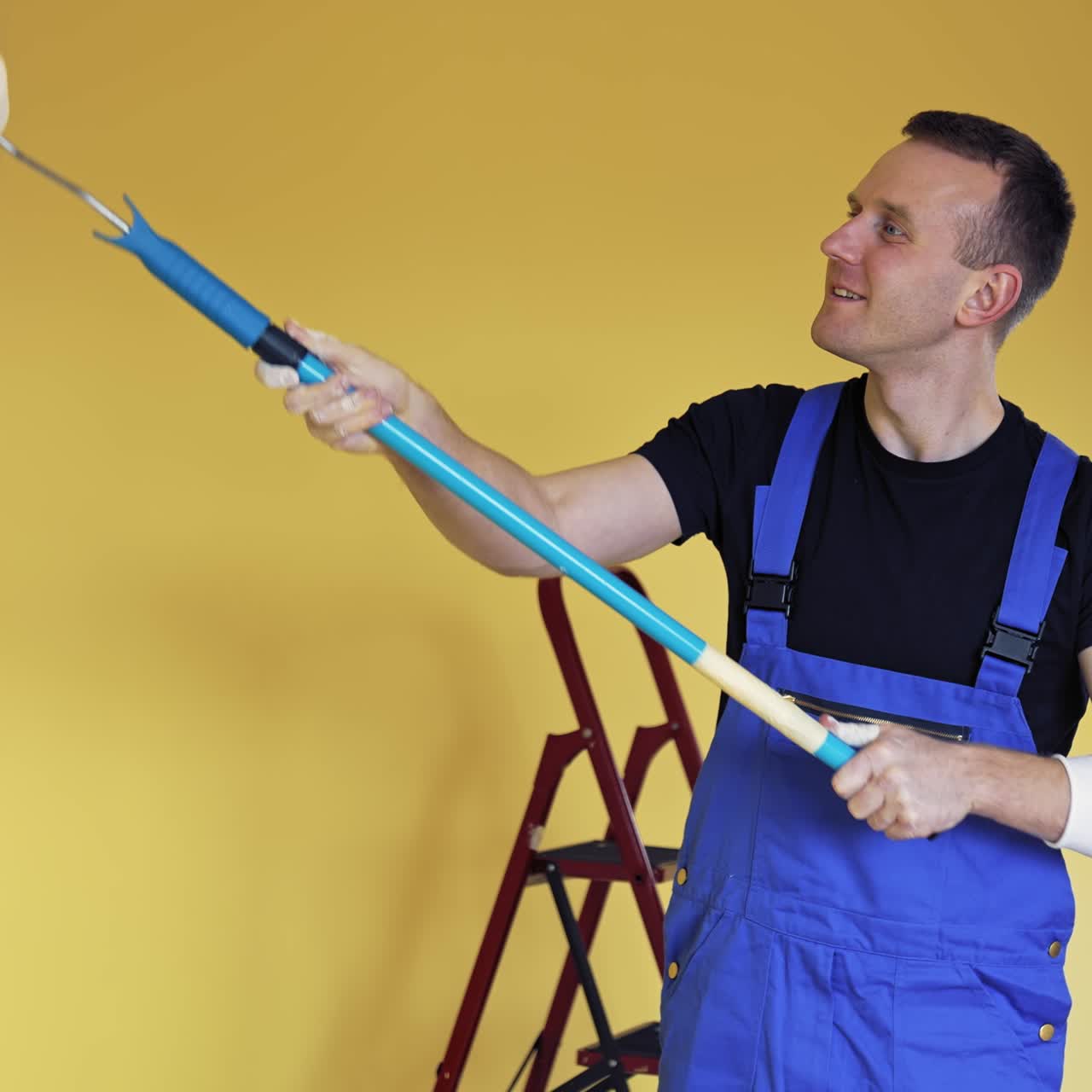 Young man doing renovation. Portrait of a painter worker doing makeover in the room. Worker in overalls paints wall with a roller brush