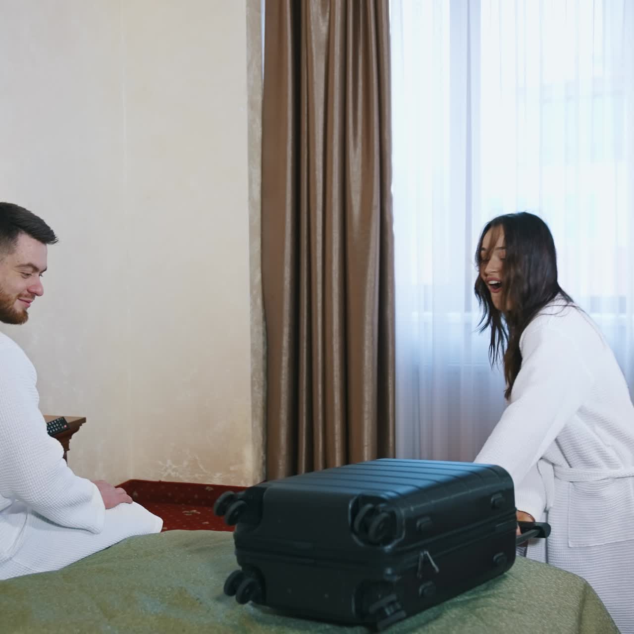 Happy woman in bathrobe with suitcase. Cheerful young female coming into the room and throwing travel bag on a bed. Young couple in dressing gowns in hotel