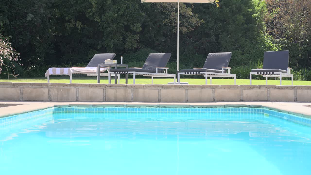 Solbeskinnet flisebelagt pool i baghaven med krusninger i vandet. Kameraet panorerer op for at afsløre en parasol, liggestole og et håndklæde.
