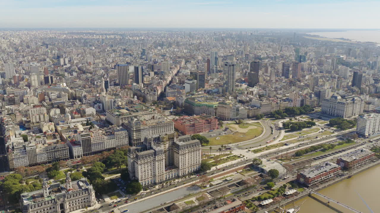 Aerial drone view over Buenos Aires shows the Casa Rosada, Plaza de Mayo, and Avenida Paseo Colón with dense city buildings stretching to the Río de la Plata under a bright sky.