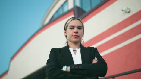 Young manager in black blazer folds hands confidently while standing in front of organization building with red and white walls and visible security camera