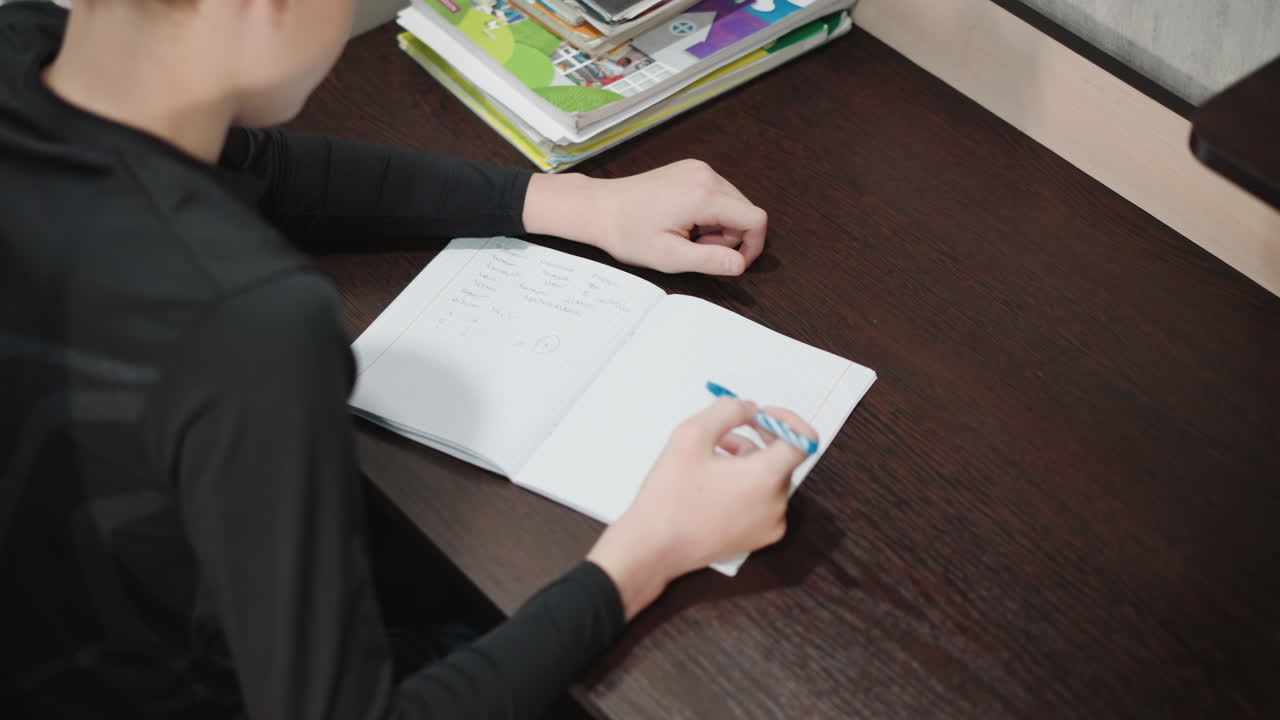 Młody chłopiec uczy się. Młody wnuczek pilnie odrabia pracę domową, z podręcznikami obok. Młody chłopiec starannie pisze w swoim zeszycie, otoczony materiałami edukacyjnymi w domu.