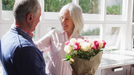 Überraschte-ältere-Frau-Mit-Blumenstrauß,-Die-Ihren-Mann-Zu-Hause-In-Der-Küche-Umarmt