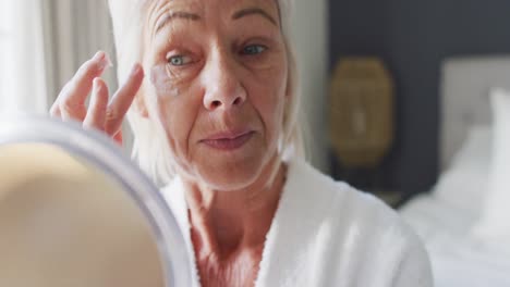 una mujer caucásica feliz en la cama en el dormitorio, mirándose en el espejo, aplicándose crema en la cara.