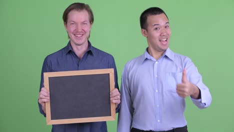 two businessman against green studio background other one holding blackboard and other showing thumbs up while smiling