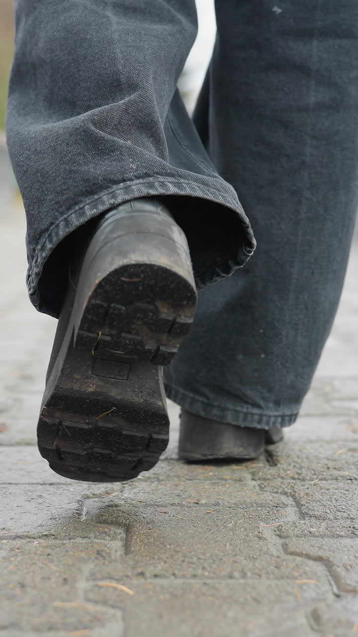 Back shot of person's legs in black trousers and black boots walking slowly on outdoor paved path during calm overcast day, capturing subtle foot movement and rhythm in peaceful urban setting