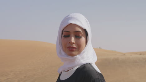 retrato de una hermosa mujer musulmana con hiyab blanco y vestido negro tradicional de pie en un desierto ventoso y sonriendo a la cámara